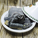 Raw dark grey-black sulfur mineral chunks displayed in a white Moroccan ceramic tagine bowl on a wooden surface, showing natural unprocessed texture with yellow sulfur veins - Variation