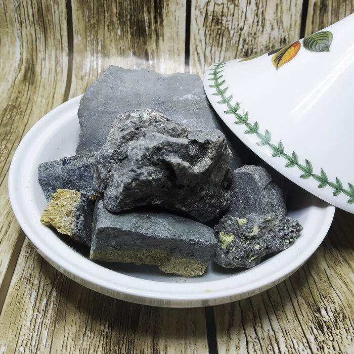 Raw dark grey-black sulfur mineral chunks displayed in a white Moroccan ceramic tagine bowl on a wooden surface, showing natural unprocessed texture with yellow sulfur veins - Variation