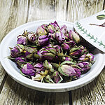 Whole dried purple-pink rose buds piled in a white ceramic bowl on a rustic wooden surface - Variation