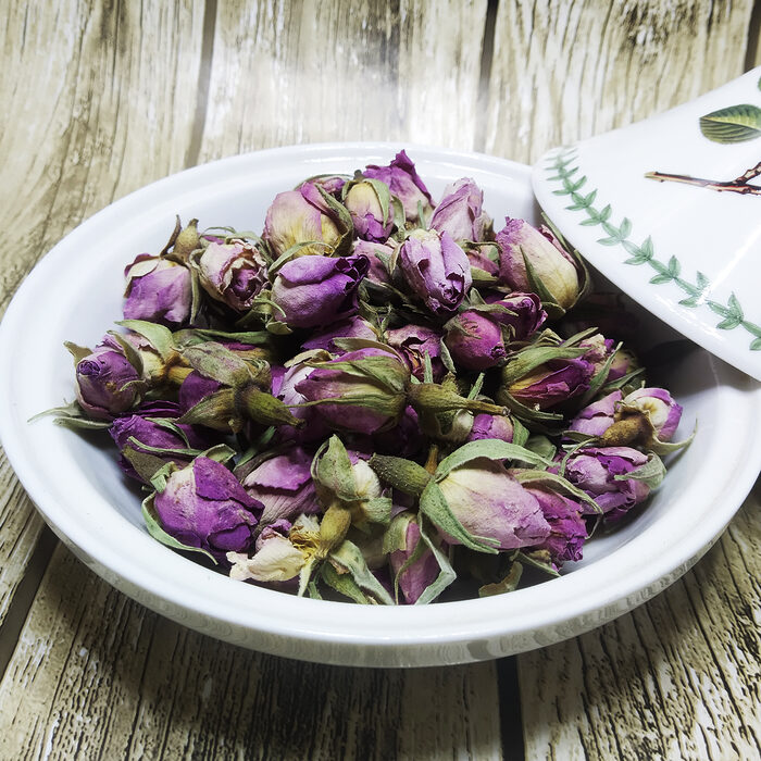 Whole dried purple-pink rose buds piled in a white ceramic bowl on a rustic wooden surface - Variation