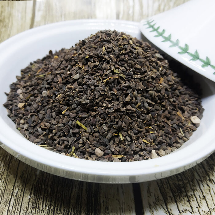 A white ceramic bowl filled with dark brown Peganum Harmala seeds on a wooden surface, with a decorative ceramic lid beside it - Variation